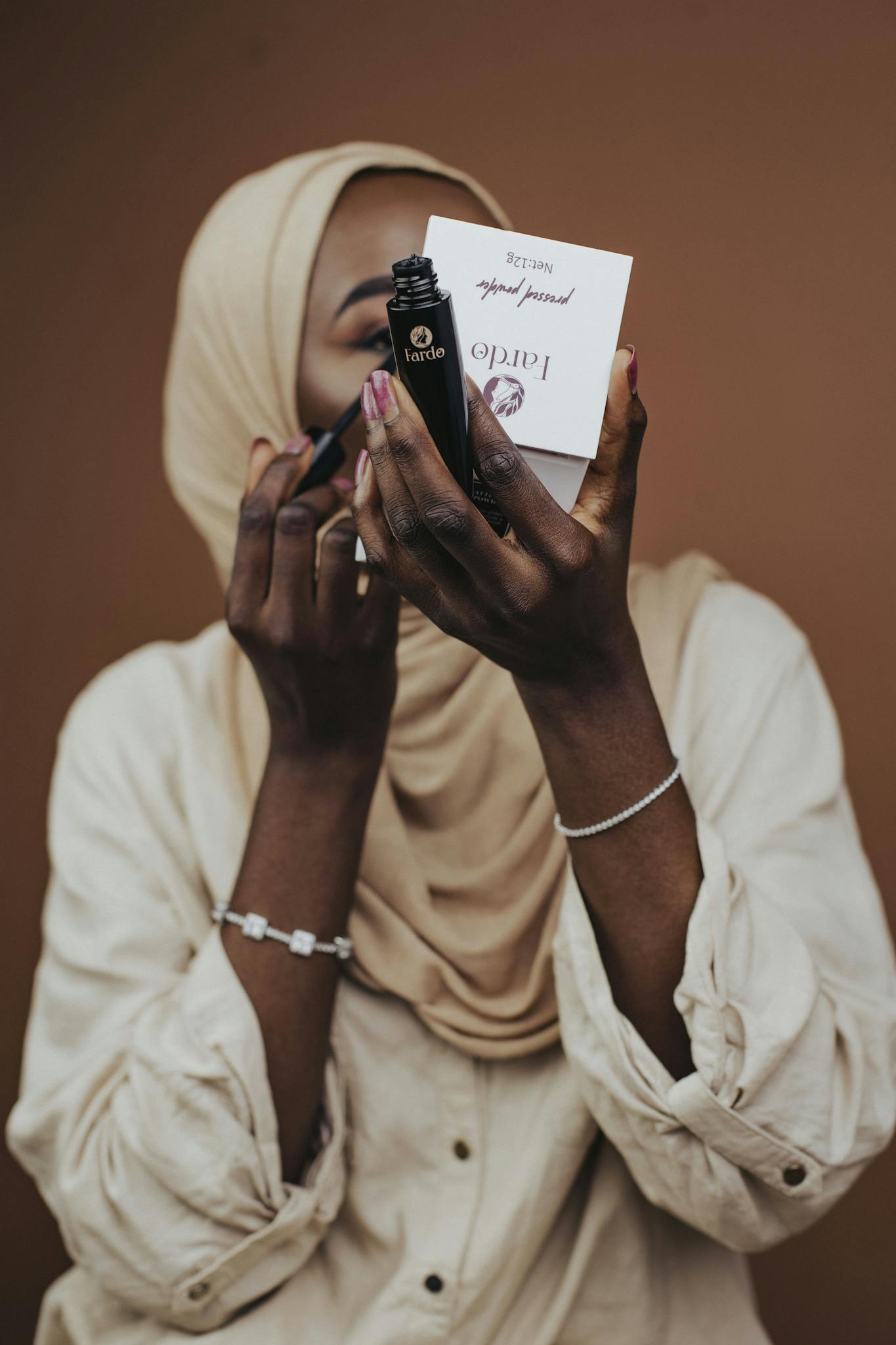 A woman in a hijab applying makeup, holding a compact mirror and lipstick.
