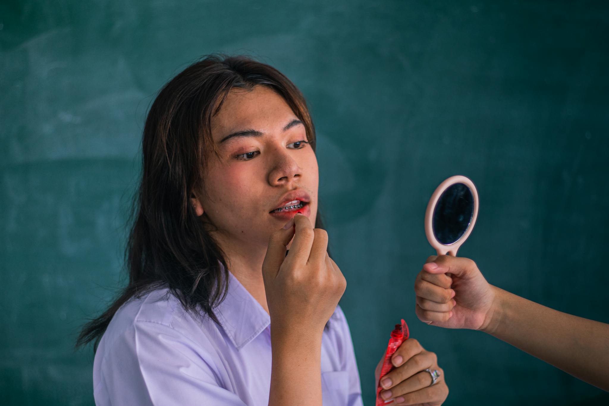A young person applies makeup with the help of a handheld mirror indoors.