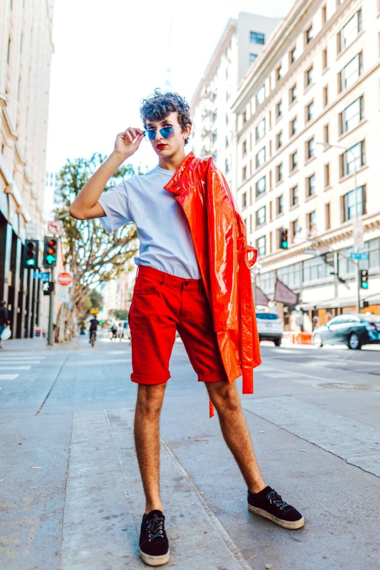Trendy young adult showcasing vibrant street fashion with a red jacket and sunglasses in a city setting.