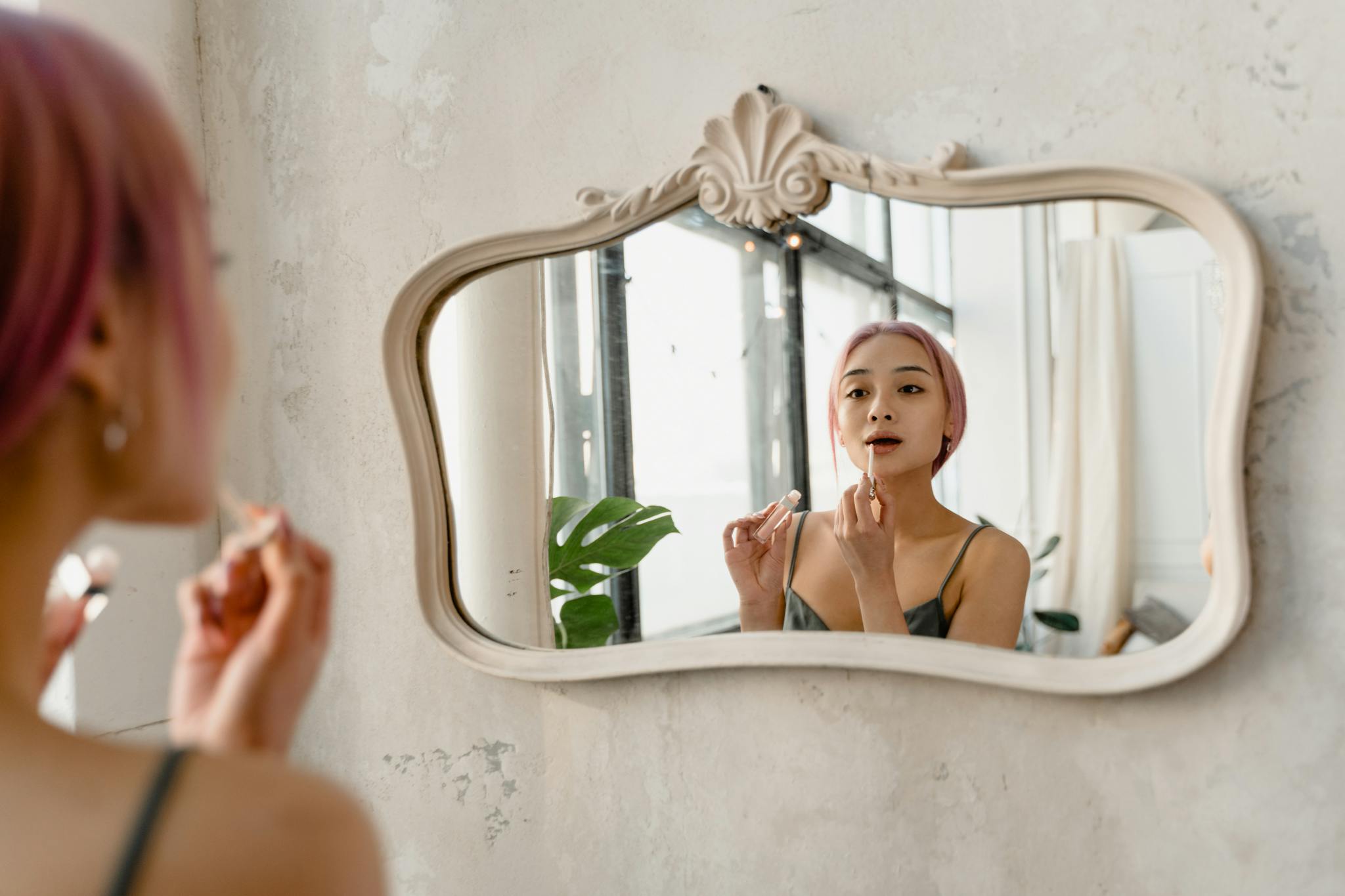 Young woman with pink hair applying makeup in front of ornate mirror indoors, embracing self-care.
